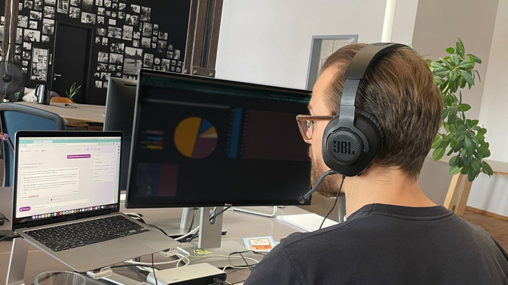 Person mit Headset vor zwei Monitoren, einer mit Diagrammen, der andere mit Text, in einem modernen Büro mit Pflanzen